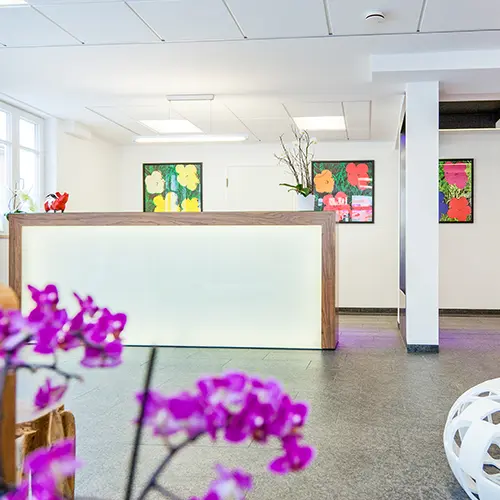 Heller, moderner Wartebereich mit rotem Sofa, Glastür, Empfangstresen und bunten Bildern an der Wand